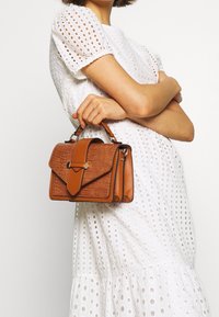 Ruskea nahkainen käsilaukku, jossa on teksturoitu krokotiilikuosi, suorakaiteen muotoinen, ylhäällä kantokahva ja läpällä suljettuna. Käytetään valkoisen mekon kanssa.