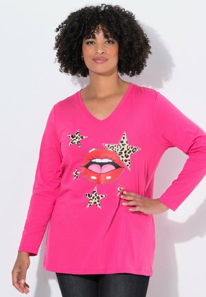 Femme aux cheveux bouclés portant un t-shirt à manches longues rose vif avec des lèvres rouges et des étoiles en imprimé léopard, posant avec une main sur la hanche.