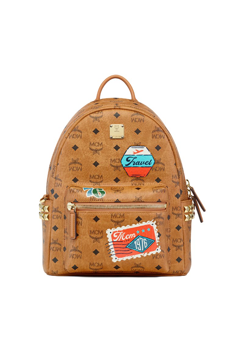 Sac à dos en cuir marron avec des logos imprimés, des écussons sur le thème du voyage, une poche zippée à l'avant et des clous pyramidaux dorés sur les accents latéraux.