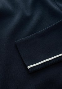 Sötétkék kötött anyag sima tapintással és fehér díszítéssel a gallér szegélyén. Hajtott gallér kialakítás jellemzi.