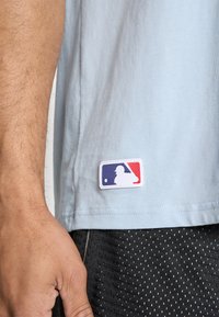Šviesiai mėlynas medvilninis marškinėliai su baltu MLB logotipo padu ir raudonomis bei mėlynomis detalėmis, dėvimi su juodais tekstūruotais šortais.