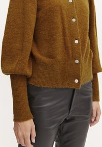 Cardigan marrone con texture, maniche lunghe e gonfiate, e polsini a coste. Presenta bottoni a forma di fiore in argento lungo la parte anteriore. Abbinato a pantaloni in pelle nera.