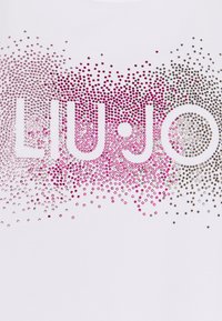 Název značky Liu Jo tvořený rozptýlenými růžovými a stříbrnými kamínky na bílém pozadí.