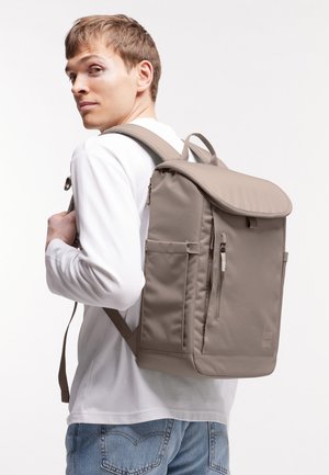 Giovane uomo che indossa uno zaino rettangolare color taupe con tasche laterali e cerniera frontale, vestito con camicia bianca e jeans blu, che guarda oltre le spalle.