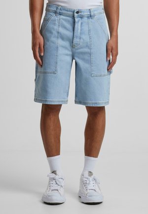 Helle blaue Denim-Shorts mit lockerem Schnitt, ausgestattet mit großen Fronttaschen, einem Reißverschluss und kontrastierenden Nähten. Kombiniert mit weißen Sneakers und Socken.