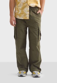 Olijfgroene cargo broek met een relaxed fit, voorzien van zijzakken en een verstelbare taille, gecombineerd met een gele bloemen blouse en sneakers.