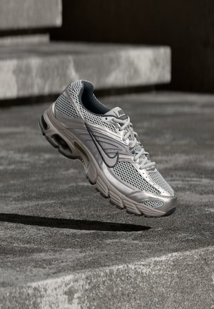Chaussure de course Nike argentée et grise suspendue en l'air au-dessus d'escaliers en béton, montrant une tige en mesh et des détails de semelle amortie.