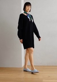 Black oversized sweater dress with long sleeves, featuring a deep V-neck. Paired with light blue loafers and a multi-colored silk scarf.