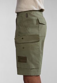 Bermudas de cargo en verde oliva con múltiples bolsillos, cierres de botón y un parche texturizado con rayas horizontales en el lado izquierdo.