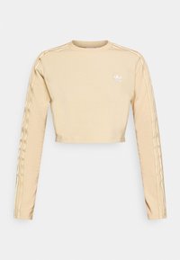 Avkortad långärmad topp i ljus beige bomull, med tre vita ränder på ärmarna och en liten broderad logotyp på bröstet. Mjuk textur.