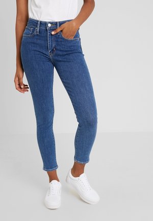 Personne portant un jean skinny bleu taille haute et des baskets blanches, debout avec une main dans la poche sur un fond uni.