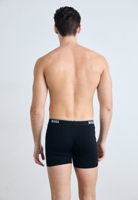 Schwarze Boxer Briefs aus glattem Stoff, mit einem weißen Bund mit "BOSS"-Branding. Sichtbare Nähte und eine enge Passform.