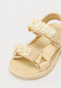 Sandale jaune clair en tissu texturé avec des embellissements floraux sur la bride et une semelle en caoutchouc durable à motifs.