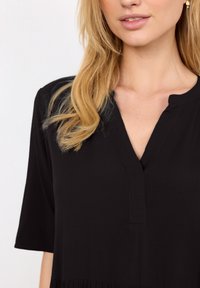 Femme blonde portant un chemisier noir à col en V avec manches jusqu'au coude et un petit détail volanté à l'ourlet, sur fond blanc.