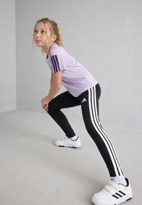 Junges Mädchen in einem lila Shirt und schwarzen Adidas-Hosen dehnt ihre Beine drinnen vor einer schlichten grauen Wand.