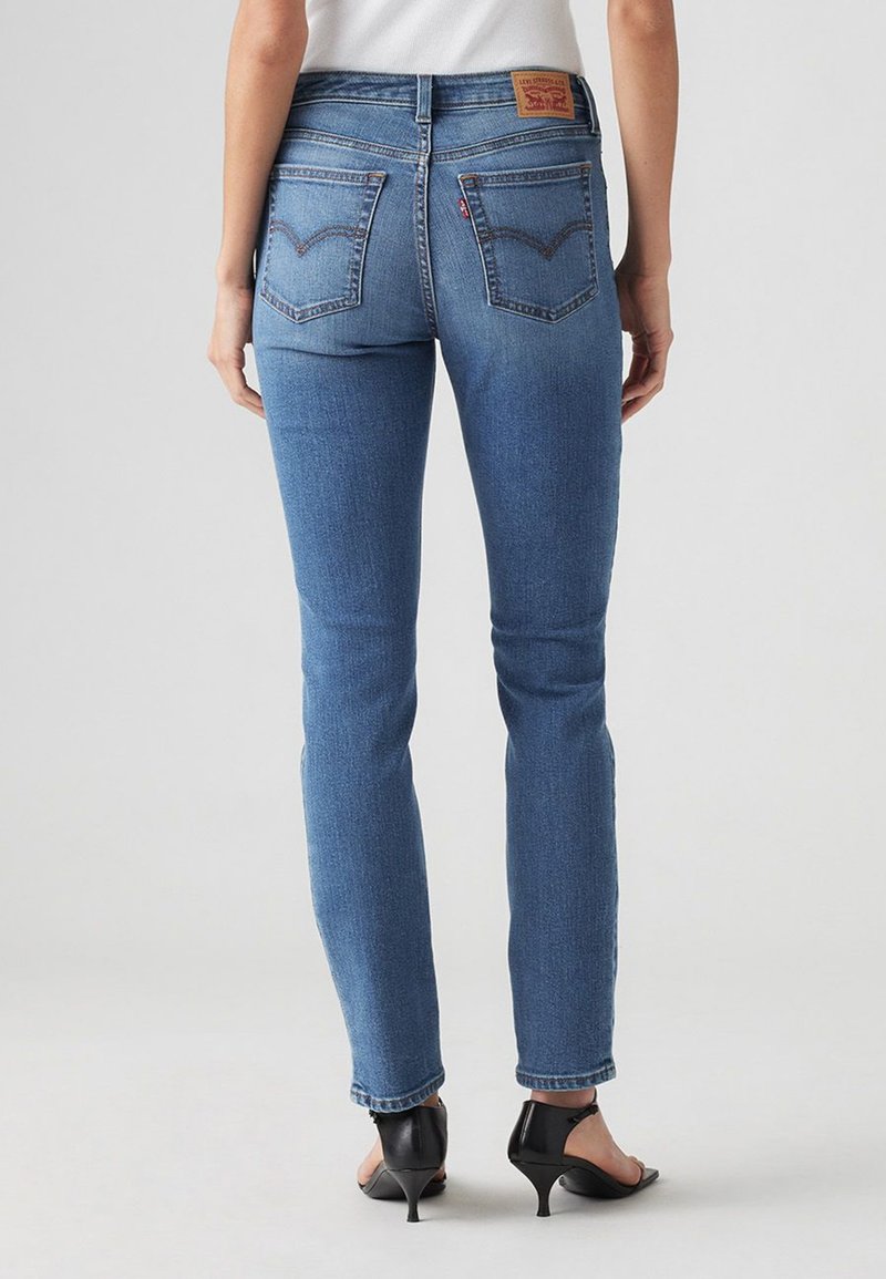 Pantalones vaqueros ajustados de mezclilla azul con cintura alta, bolsillos traseros y costuras distintivas. Combinados con tacones abiertos negros.