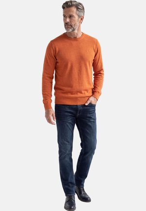 Mężczyzna z siwymi włosami i brodą, ubrany w pomarańczowy sweter z długim rękawem, ciemnoniebieskie dżinsy i czarne skórzane buty, stoi z jedną ręką w kieszeni.