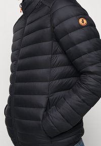 Czarna kurtka puchowa z poziomym pikowaniem, stójką i pomarańczową naszywką z logo na ramieniu. Zamykane na zamek kieszenie po bokach.