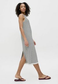 Vestido de rayas sin mangas en tejido de punto negro y blanco, de largo hasta la mitad de la pantorrilla con aberturas laterales. Combinado con chanclas planas de color púrpura oscuro.