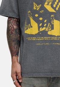 Szara grafika koszulka z żółtym nadrukiem przedstawiającym kości, motyle i kwiatowe wzory. Luźny krój, krótki rękaw i okrągły dekolt.