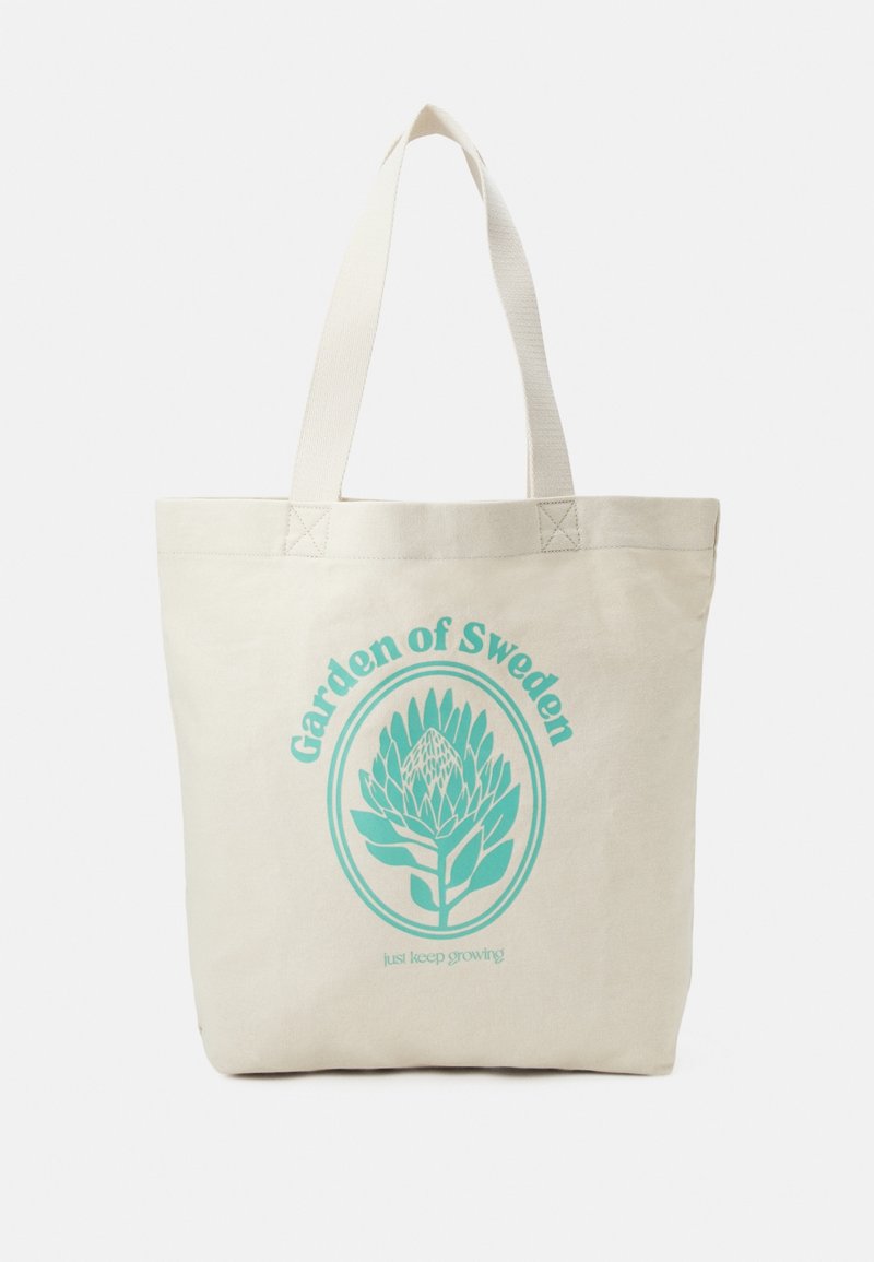 Borsa tote in canvas di colore naturale con un design grafico turchese che presenta il testo "Giardino di Svezia" e un'illustrazione di pianta.