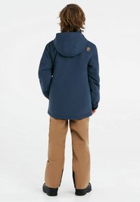 Giacca impermeabile navy con cappuccio, polsini elasticizzati e logo sul retro, abbinata a pantaloni da neve marrone chiaro e stivali neri.