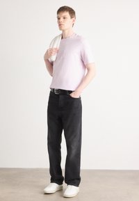 Lyse lilla bomulds-t-shirt med broderet logo, parret med sorte jeans og hvide sneakers. Modellen holder et hvidt håndklæde over skulderen.