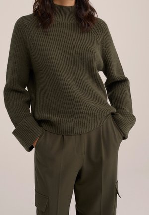 Žena v tmavě zeleném žebrovaném svetru s ohrnutými rukávy a volných cargo kalhotách v odpovídající barvě, ruce v kapsách, neutrální pozadí.