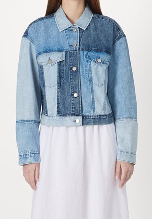 Woman wearing a color-blocked denim jacket with silver buttons over a white dress, standing against a plain white background.