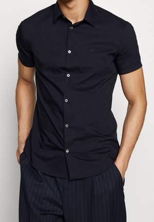 Homme portant une chemise noire cintrée à manches courtes avec un petit logo sur la poitrine, associée à un pantalon sombre à fines rayures, mains dans les poches.