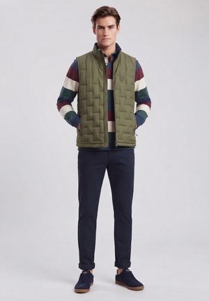 Hombre con chaleco acolchado verde sobre camisa de rayas de manga larga, pantalones oscuros remangados y zapatillas azul marino, de pie con las manos en los bolsillos del chaleco.