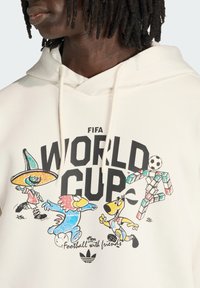 Φούτερ με κουκούλα που έχει κείμενο FIFA World Cup και πολύχρωμους κινουμένων σχεδίων μασκότ να παίζουν ποδόσφαιρο, με τη φράση «ποδόσφαιρο με φίλους» από κάτω.