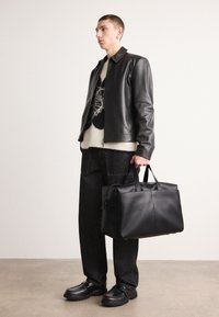 Borsa a tote nera in pelle con design strutturato, forma rettangolare, doppi manici e superficie liscia, tenuta da una persona in abbigliamento scuro.