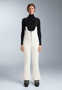 Nő fekete hosszú ujjú garbóban, fehér kötött kantáros nadrágban cipzárral az elején és fekete csattal ellátott csizmában, szürke háttér előtt állva.