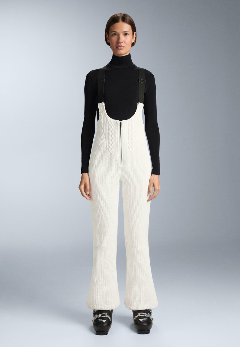 Nő fekete hosszú ujjú garbóban, fehér kötött kantáros nadrágban cipzárral az elején és fekete csattal ellátott csizmában, szürke háttér előtt állva.