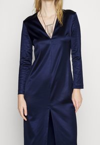 Navy satin dress with a V-neck, long sleeves, and a front slit. Worn with a silver necklace featuring multiple chain accents.