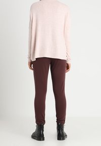 Übergröße rosafarbener Pullover mit strukturierter Oberfläche, kombiniert mit schmal geschnittenen dunkelbraunen Hosen und schwarzen Stiefeletten. Ansicht von hinten, minimaler Hintergrund.