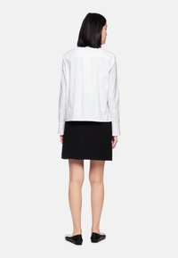 White long-sleeve shirt, relaxed fit, buttoned cuff, black textured skirt, above knee length, flat black shoes. Model shown from the back.
