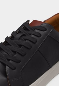 Μαύρο δερμάτινο αθλητικό παπούτσι με μαύρα κορδόνια, καφέ γλώσσα και λευκή λαστιχένια σόλα σε λευκό φόντο.