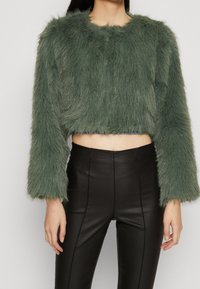 Beskåret grøn faux fur jakke med lange ærmer, der har en struktureret overflade. Sammen med højtaljede sorte læderbukser.