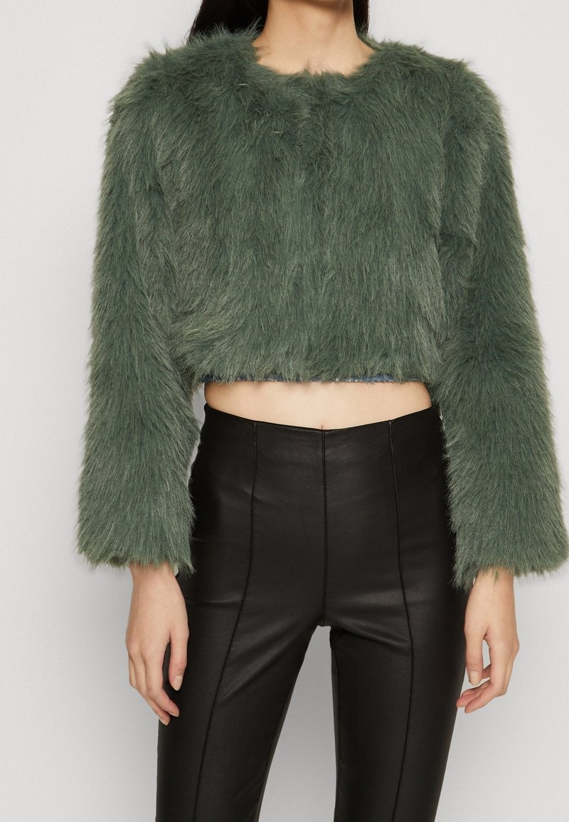 Beskåret grøn faux fur jakke med lange ærmer, der har en struktureret overflade. Sammen med højtaljede sorte læderbukser.