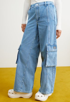 Pantalons cargo en denim bleu clair avec des jambes larges, dotés de poches latérales et d'une étiquette patch. Portés avec des sabots blancs sur une surface jaune.