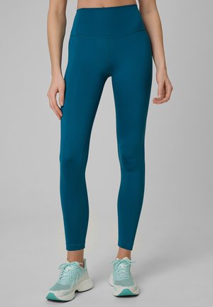 Mujer con mallas de cintura alta en color verde azulado y zapatillas deportivas azul claro, de pie frente a un fondo gris liso.