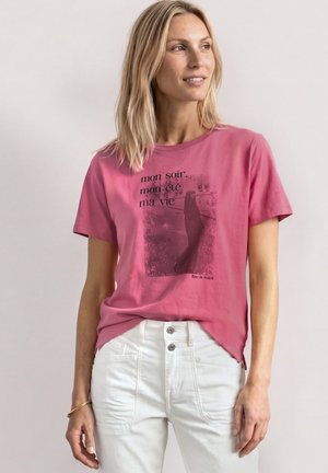 Kvinde med skulderlangt blondt hår iført en pink t-shirt med grafik og hvide bukser, der ser lidt mod højre med et smil.