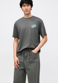 T-shirt cinzenta em algodão com mangas curtas e gola redonda, com estampa gráfica em tom verde-azulado no peito esquerdo. Combinada com calças cinzentas com cordão.