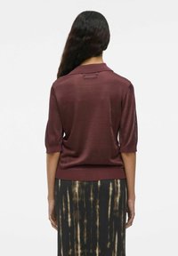 Donna con lunghi capelli scuri che indossa un top a maniche corte color bordeaux e una gonna nera con motivi a strisce verticali di tie-dye beige, in piedi su uno sfondo bianco.