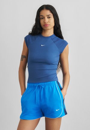 Jeune femme aux longs cheveux foncés porte un haut bleu ajusté Nike et un short de sport Nike bleu vif, debout les mains dans les poches.