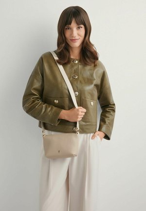 Kobieta o brązowych włosach, ubrana w oliwkowozieloną skórzaną kurtkę i beżowe spodnie, trzymająca beżową torebkę na ramię na jednolitym tle.