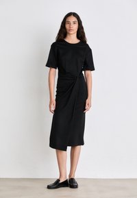 Abito nero fino al ginocchio realizzato in tessuto morbido, con scollatura rotonda, maniche corte e dettaglio intrecciato in vita. Scarpe nere basse completano l'outfit.