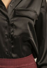 Blouse en satin noir avec une poche avant, fermeture à boutons et une texture lisse, associée à une ceinture à rayures bordeaux.